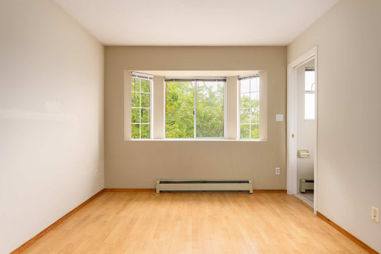 Empty room with light wood flooring, beige walls, a large bay window facing green trees, and a doorway leading to a small room with a toilet visible.
