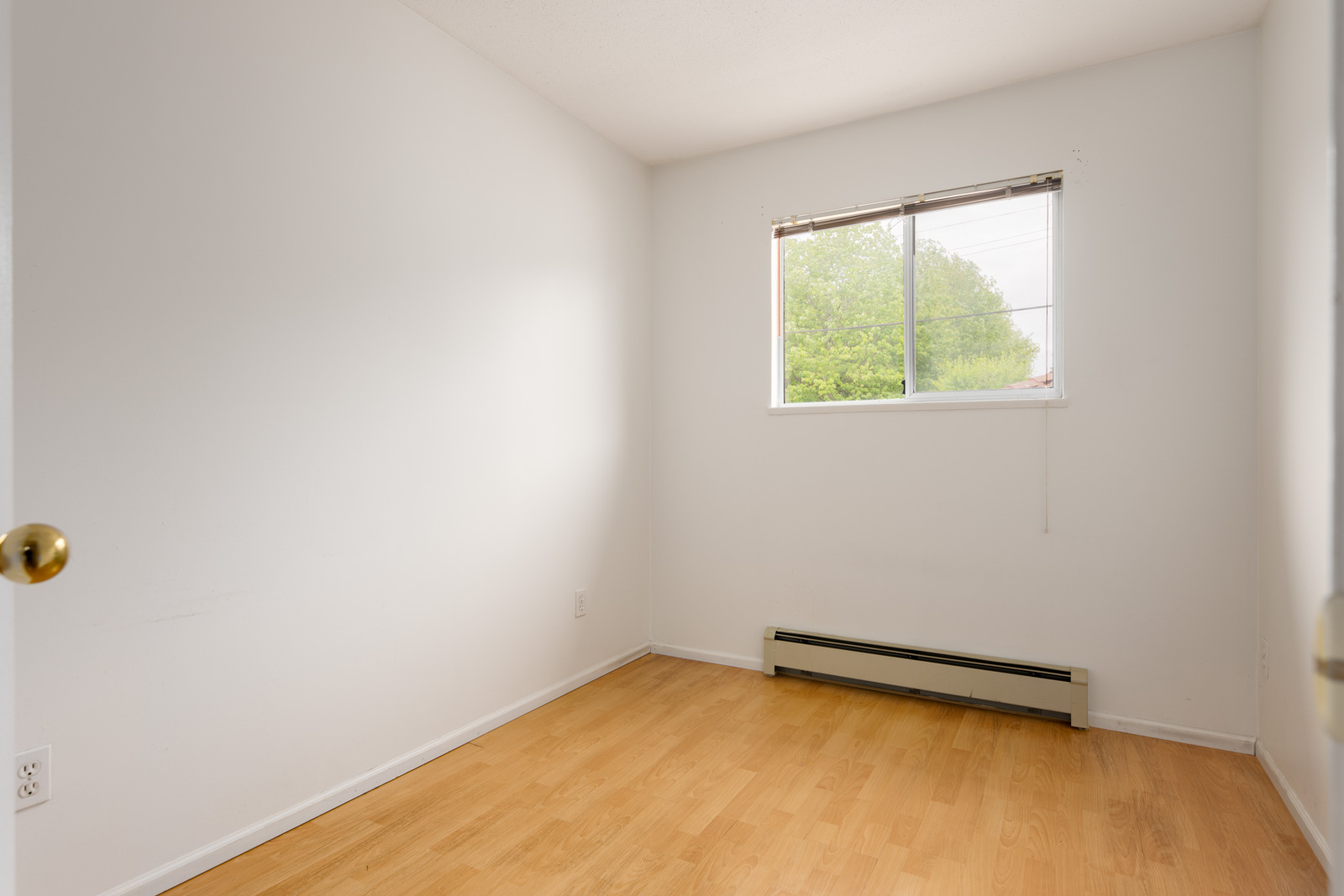Small empty room with white walls, light wood flooring, a baseboard heater, and a single window letting in natural light.