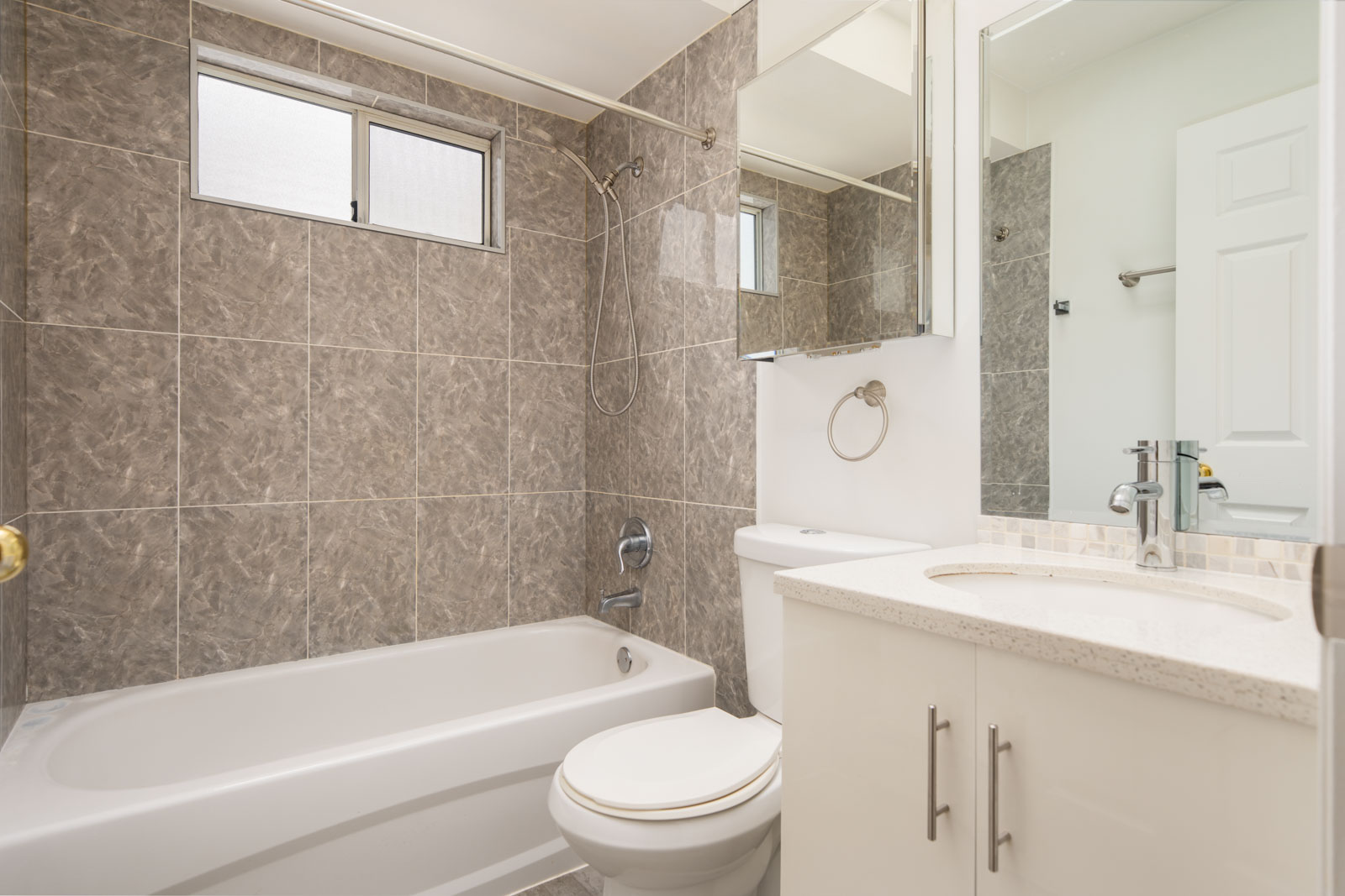 Modern bathroom with gray tiled walls, a white bathtub-shower combo, toilet, white vanity with sink, and mirrored cabinet above the sink.