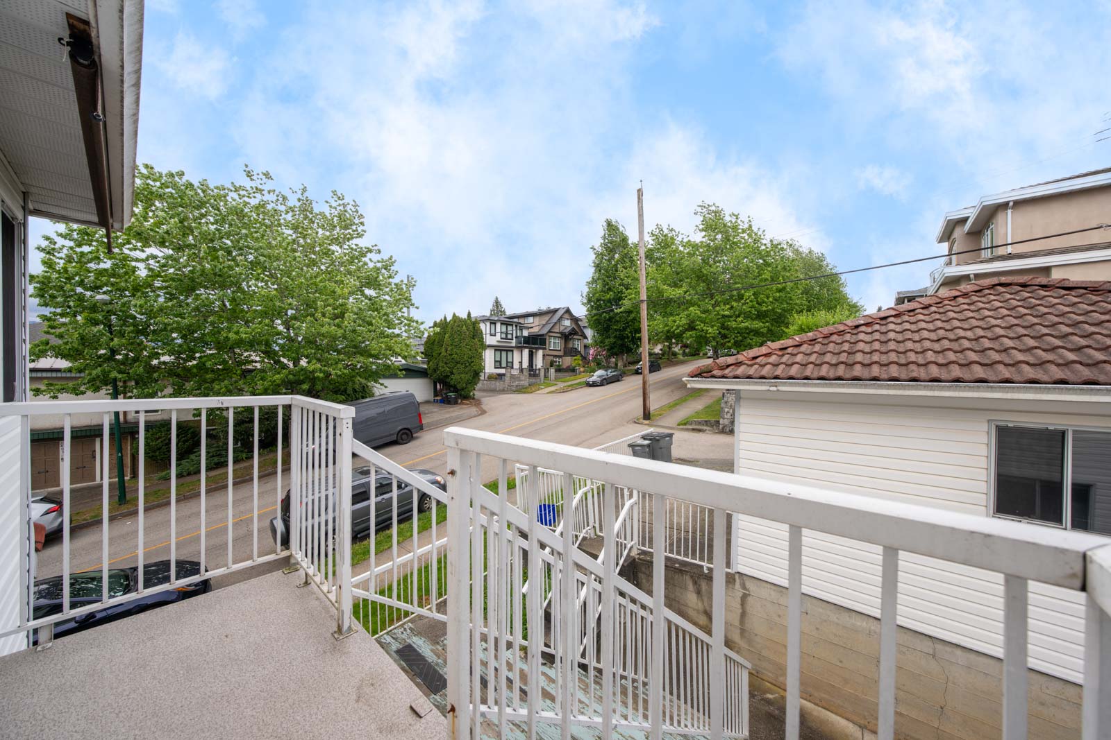 View from a balcony showing metal railings, stairs, nearby houses, a street with a parked van, and trees under a partly cloudy sky.