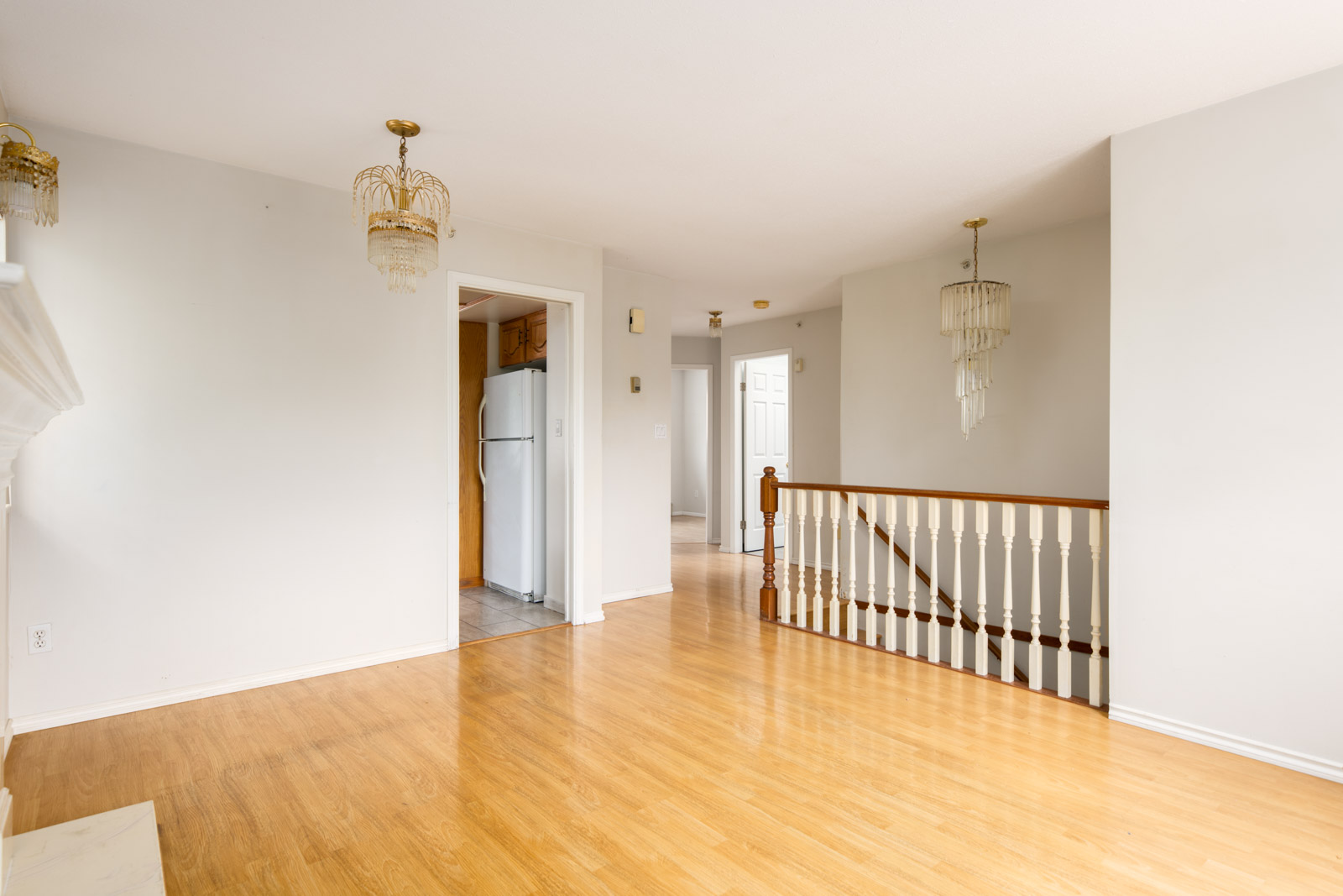 Unfurnished room with light wood flooring, white walls, elegant chandeliers, a staircase with wooden railing, and a view into a kitchen with a refrigerator.