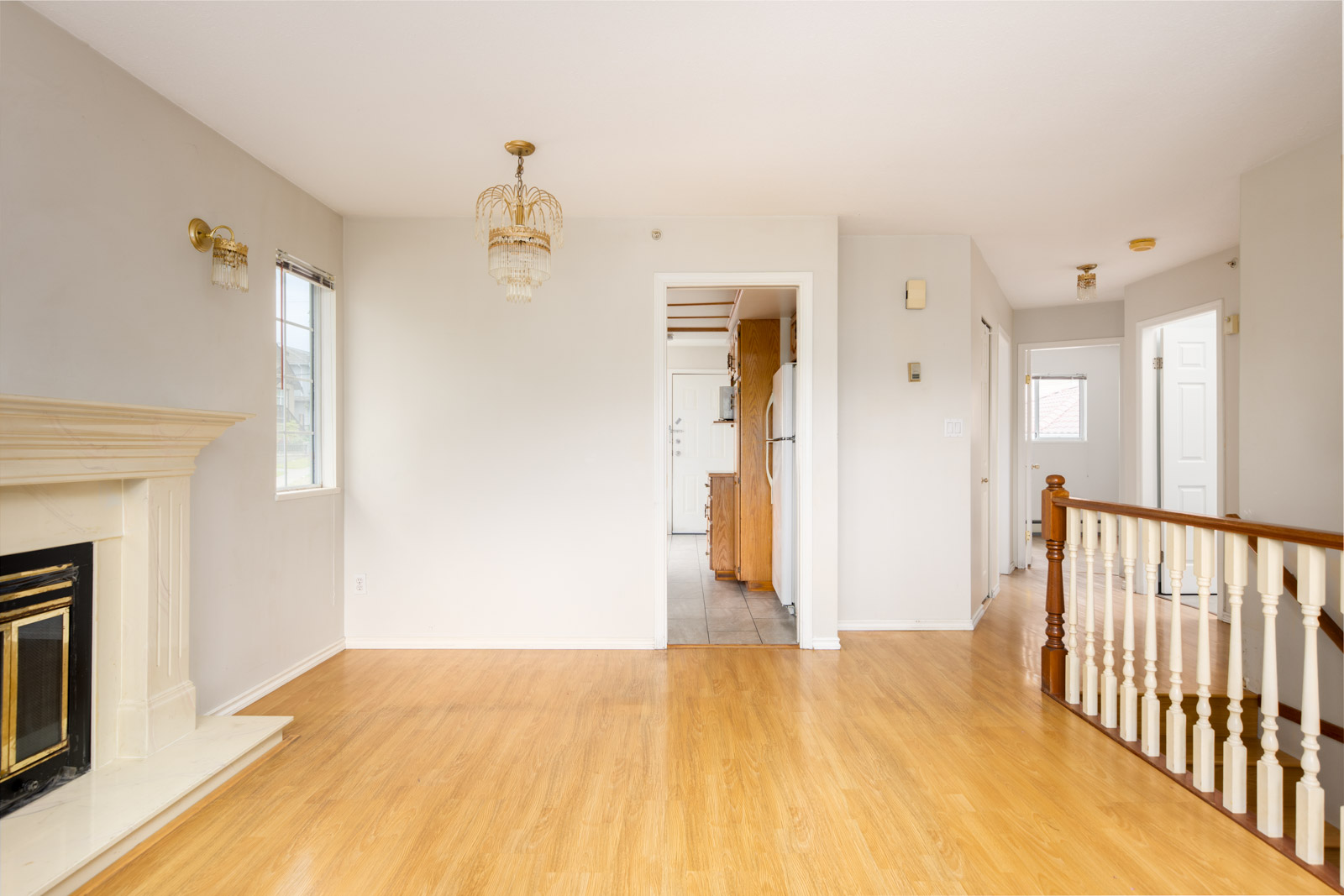 Bright, empty room with light wood flooring, a white fireplace, and a chandelier, leading to a tiled kitchen area and a hallway with white doors and a wooden banister.