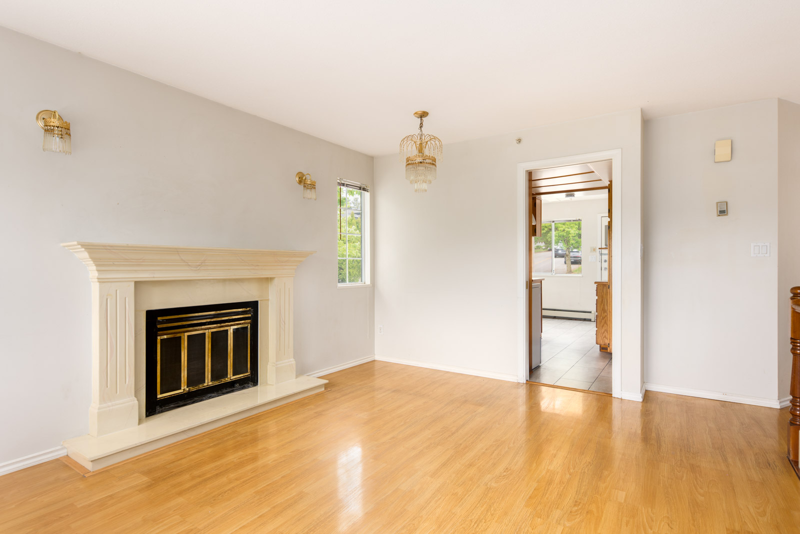 Unfurnished living room with light wood floors, white walls, a decorative fireplace, wall sconces, and a view into a kitchen through an open doorway.