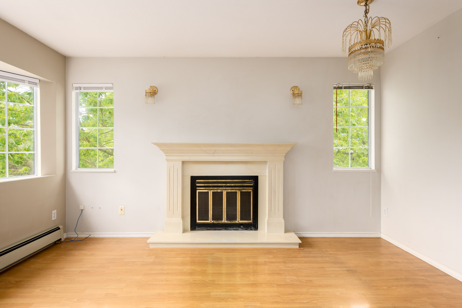 Empty room with light-colored walls, two windows, a white fireplace with a brass-trimmed insert, wooden floor, and a chandelier hanging from the ceiling.