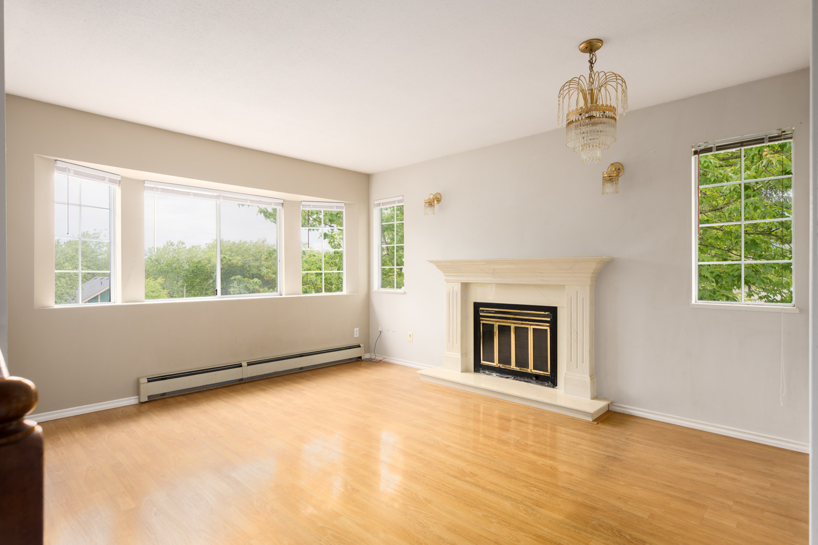 Empty living room with light wood floors, beige walls, a white fireplace, large bay window, two side windows, and a chandelier. Green trees visible outside.