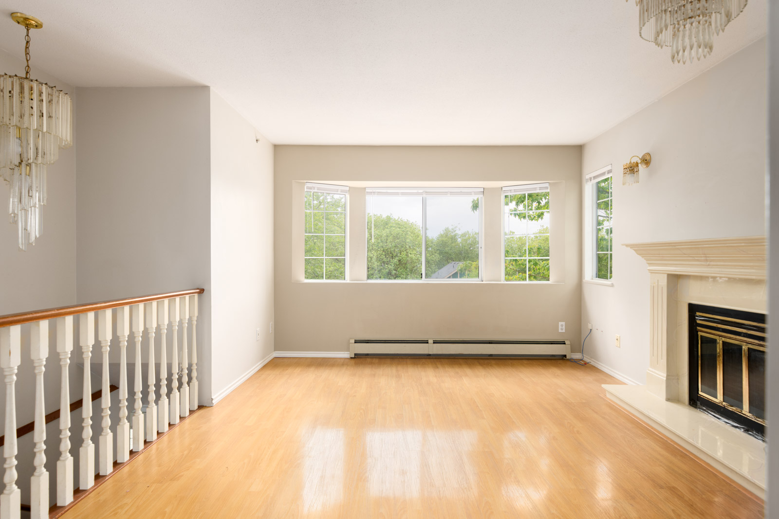 Bright, empty living room with wood flooring, large windows overlooking trees, a white staircase railing, and a fireplace with a gold-trimmed cover.