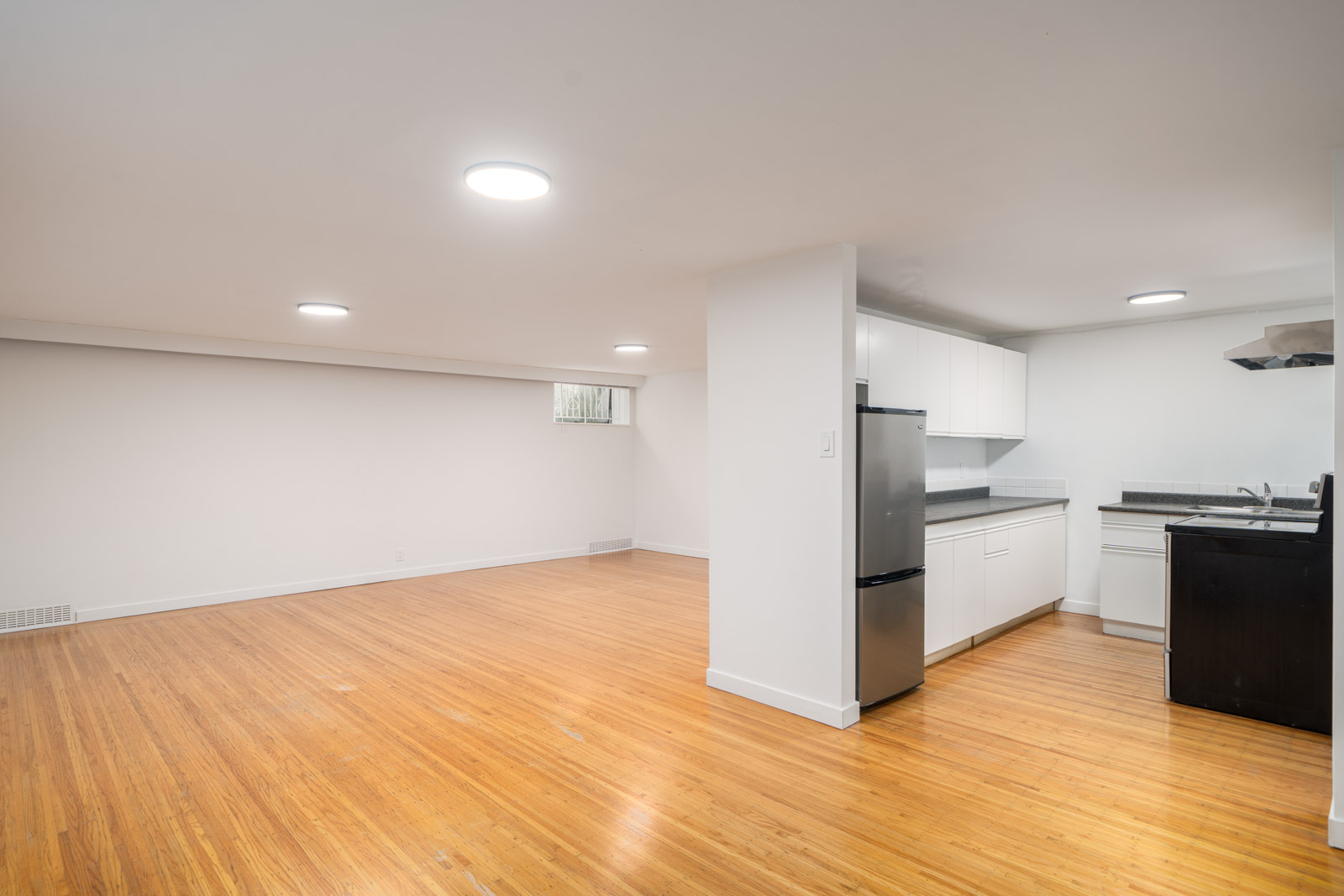 Unfurnished basement apartment with wood flooring, white walls, recessed lighting, and an open kitchen featuring white cabinets and stainless steel appliances.