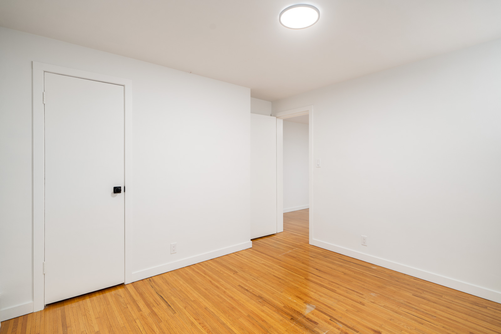 Empty room with white walls, a closed white door, an open doorway, and light wood flooring under a round ceiling light.
