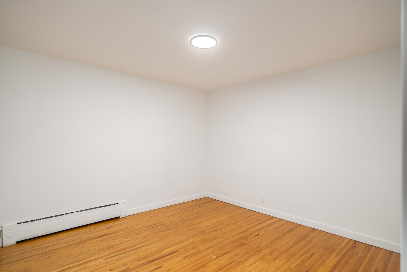 Empty room with white walls, a ceiling light, wooden floor, and a baseboard heater along one wall.