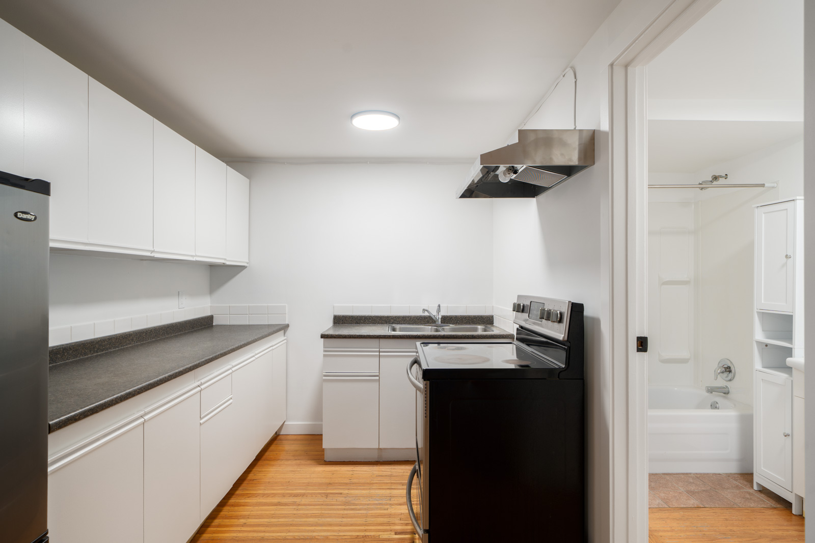 A modern kitchen with white cabinets, black countertops, stainless steel appliances, and an adjacent bathroom visible through an open door.