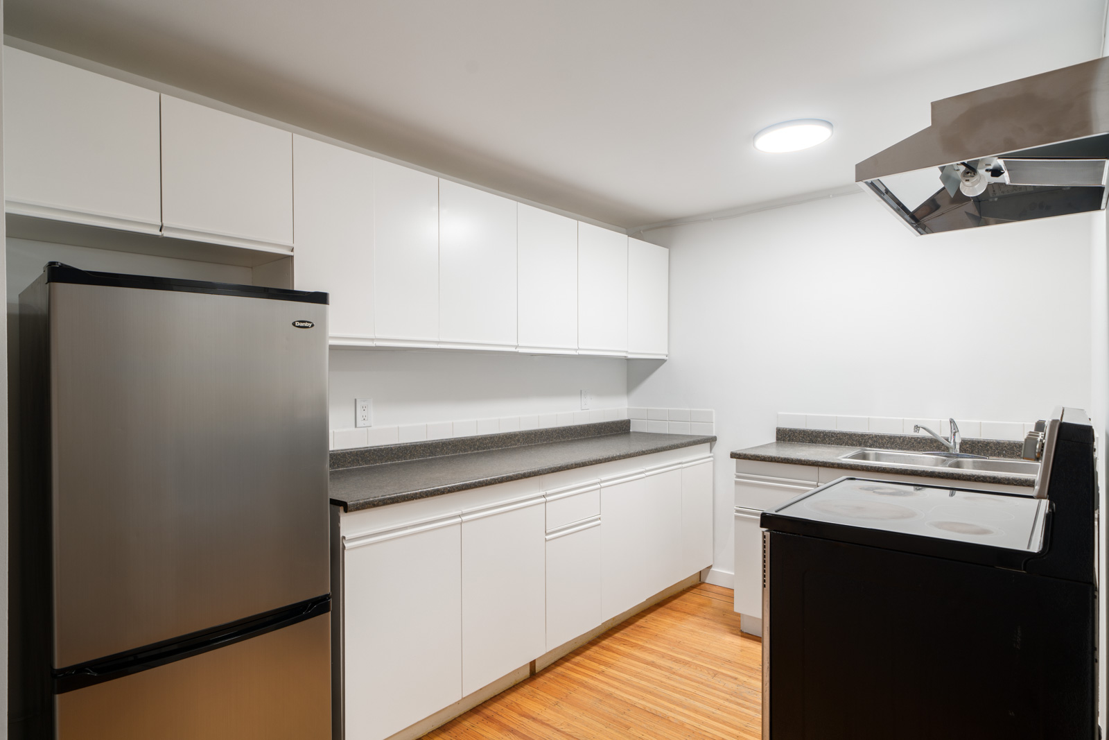 Modern kitchen with white cabinets, stainless steel refrigerator, electric stove, sink, and wooden flooring under bright ceiling light.