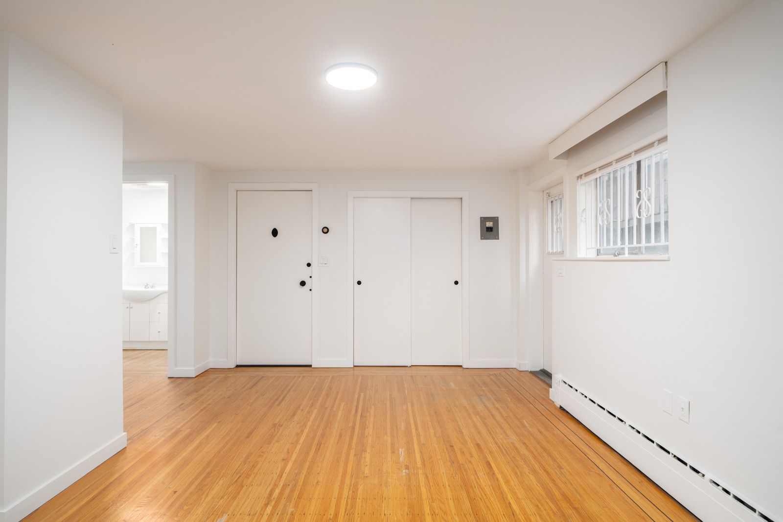 Bright empty room with wooden floors, white walls, closed white doors, large window with metal bars, and baseboard heating along the wall.