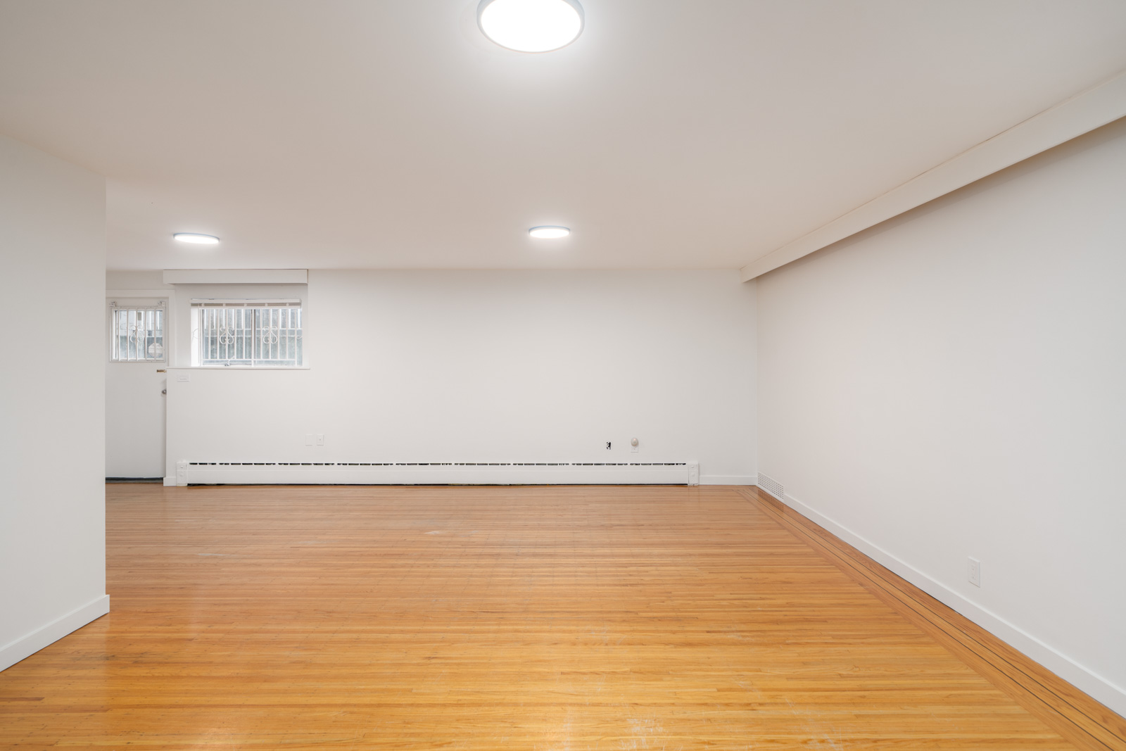 An empty room with white walls, two ceiling lights, a wooden floor, a window with bars, and a door on the left side.