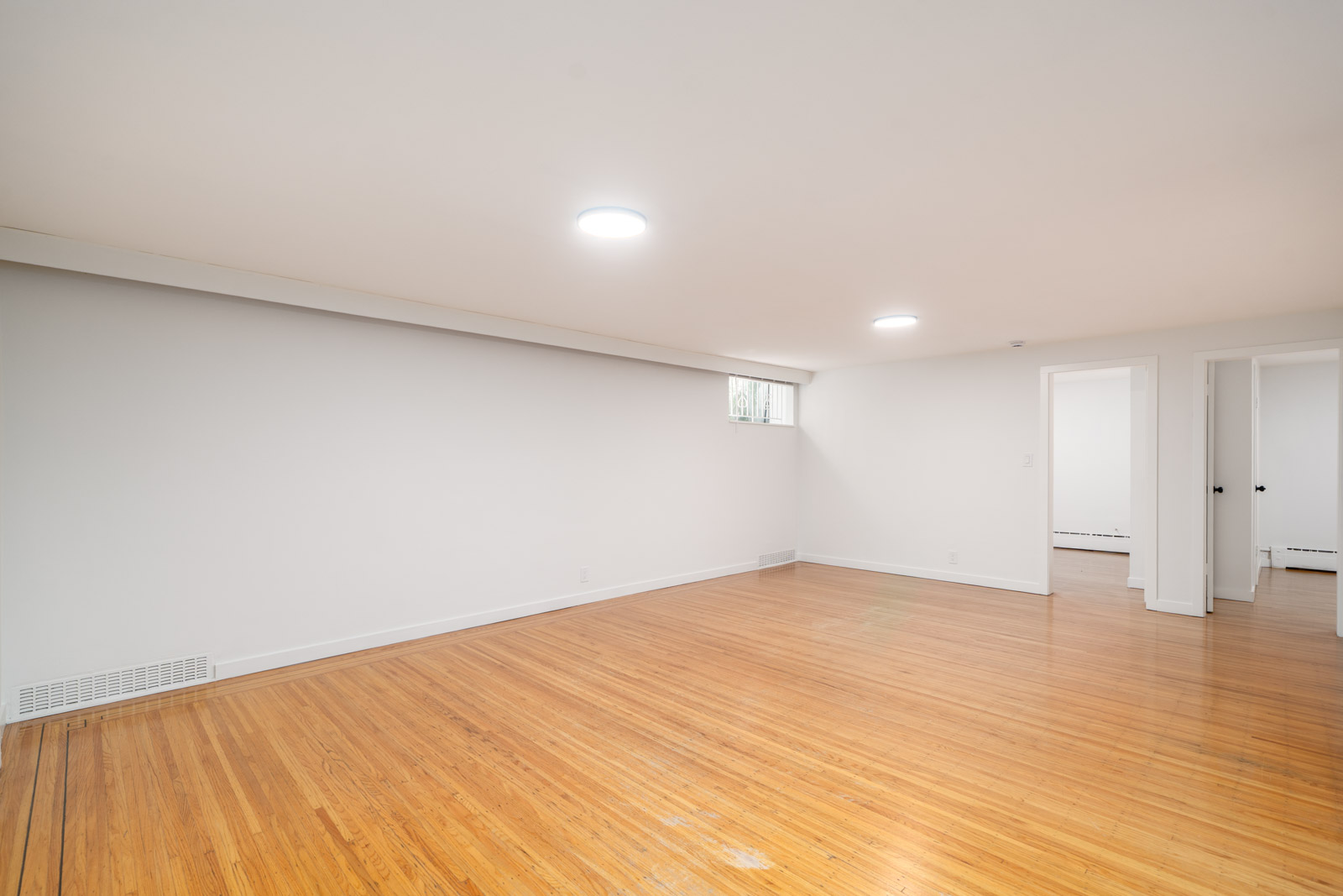 A spacious, empty room with light-colored walls, hardwood floors, two ceiling lights, a small window, and doorways leading to adjacent rooms.