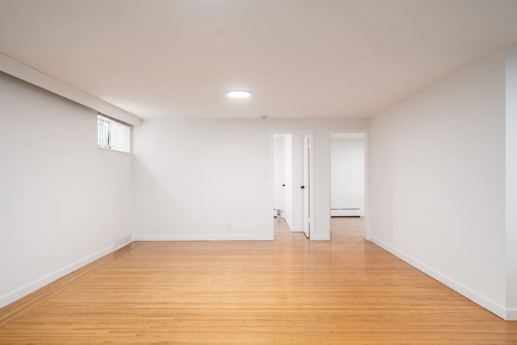 Unfurnished basement room with white walls, wood flooring, a small window near the ceiling, and two open doorways leading to other rooms.
