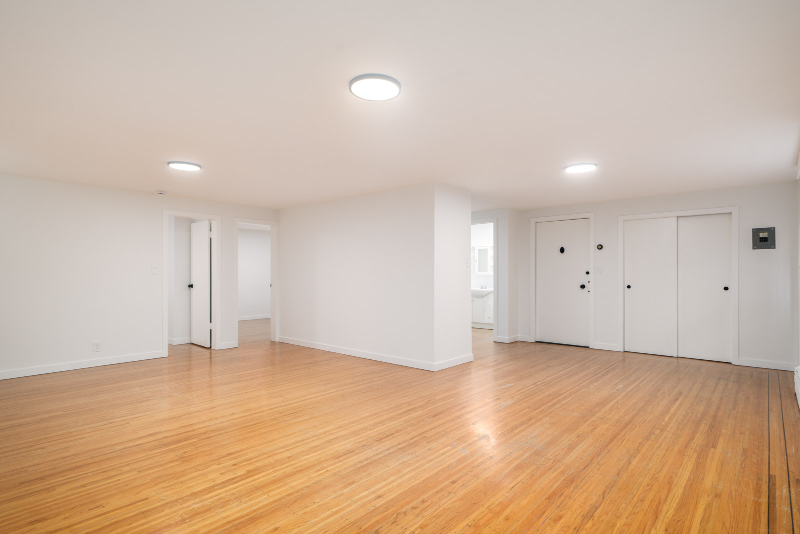 Spacious, empty room with light-colored walls, wood flooring, and ceiling lights. Multiple doors are visible, including closets and entrances to other rooms.