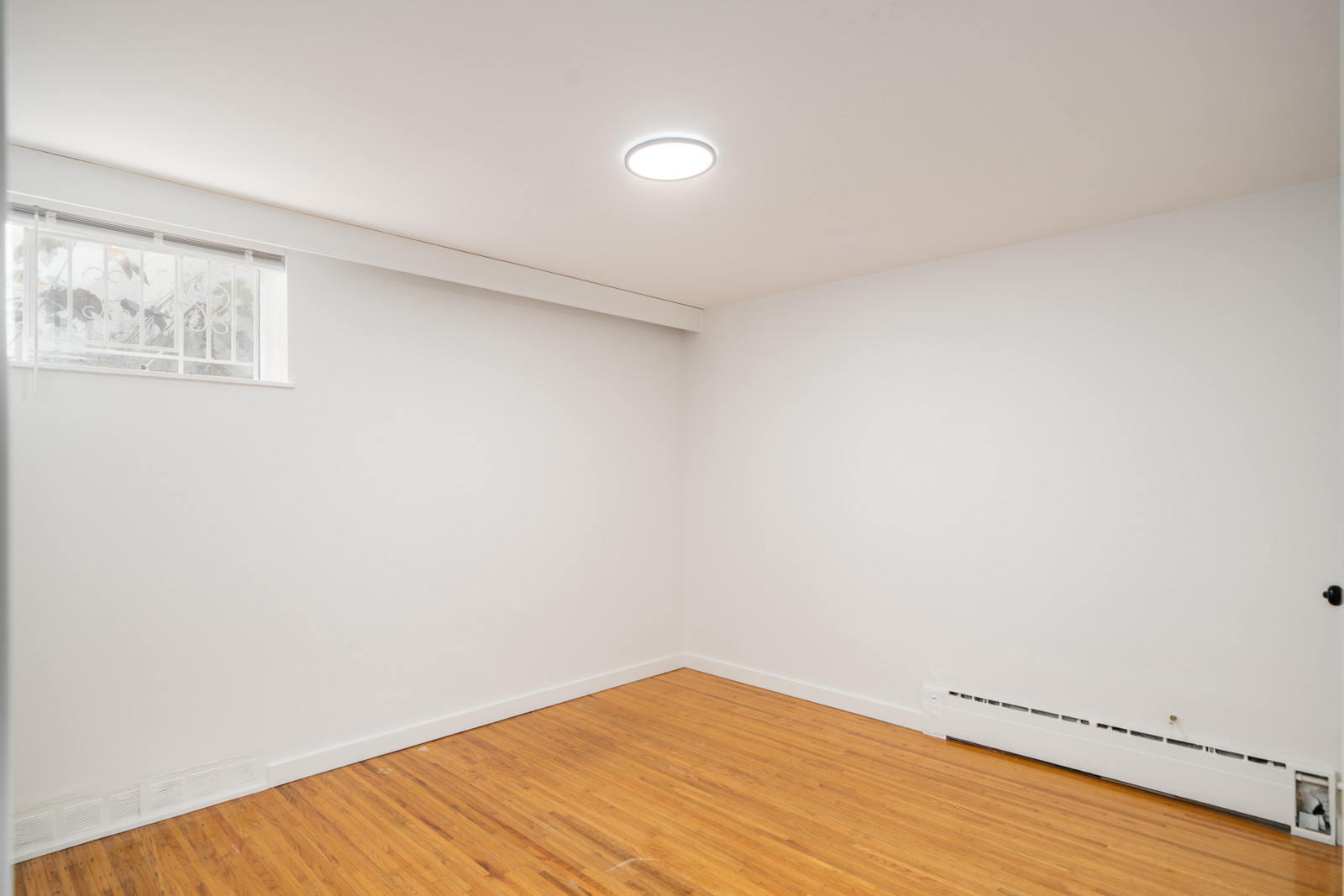 Empty room with white walls, a small window, wooden floor, ceiling light, and a white baseboard heater along one wall.