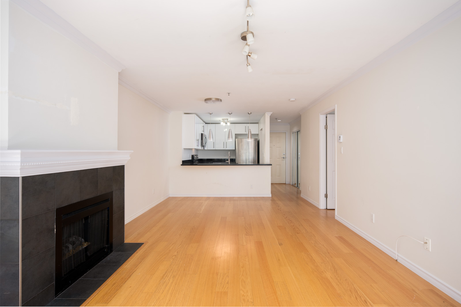 Empty living room with hardwood floors, a black fireplace on the left, and an open view into a modern kitchen with white cabinets and stainless steel appliances.