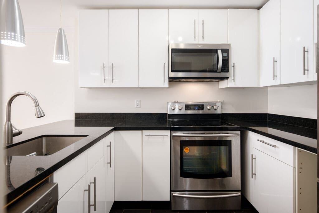 Modern kitchen with white cabinets, black countertops, stainless steel appliances including an oven and microwave, and a sink with a gooseneck faucet under pendant lights.