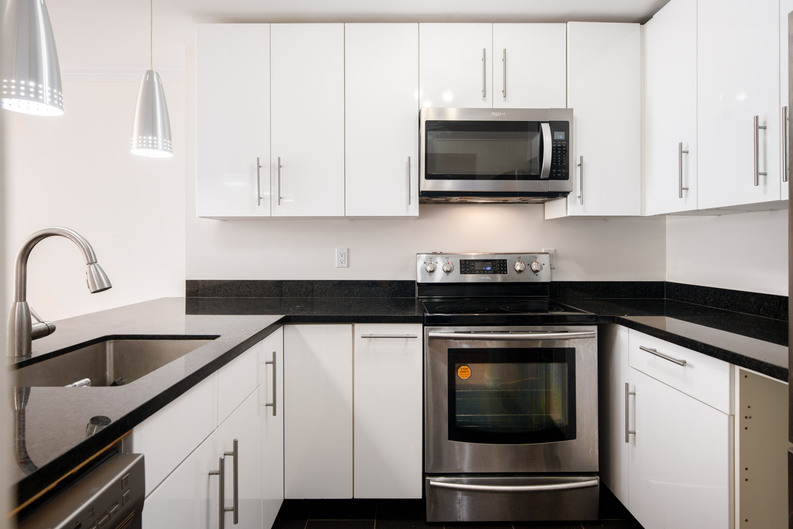 Modern kitchen with white cabinets, black countertops, stainless steel appliances including an oven and microwave, and a sink with a gooseneck faucet under pendant lights.
