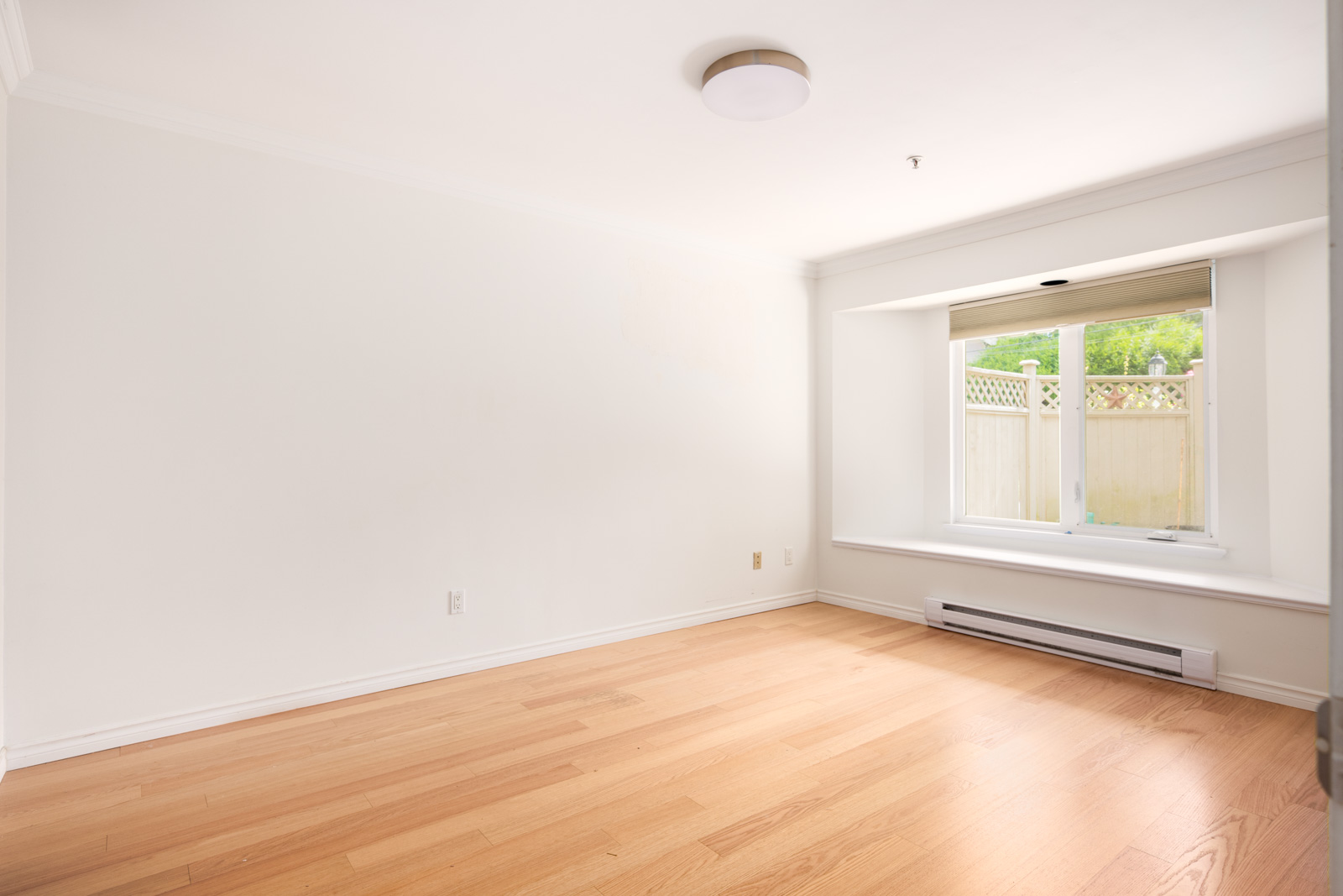 Empty room with light wood flooring, white walls, a round ceiling light, a baseboard heater, and a large window with a view of a fenced outdoor area and greenery.