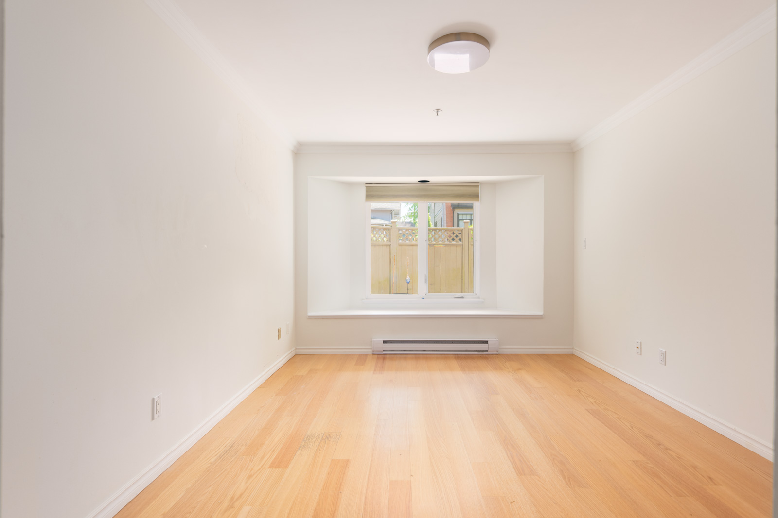Empty room with light-colored wooden flooring, white walls, a ceiling light, and a window with a view of a wooden fence and greenery outside.