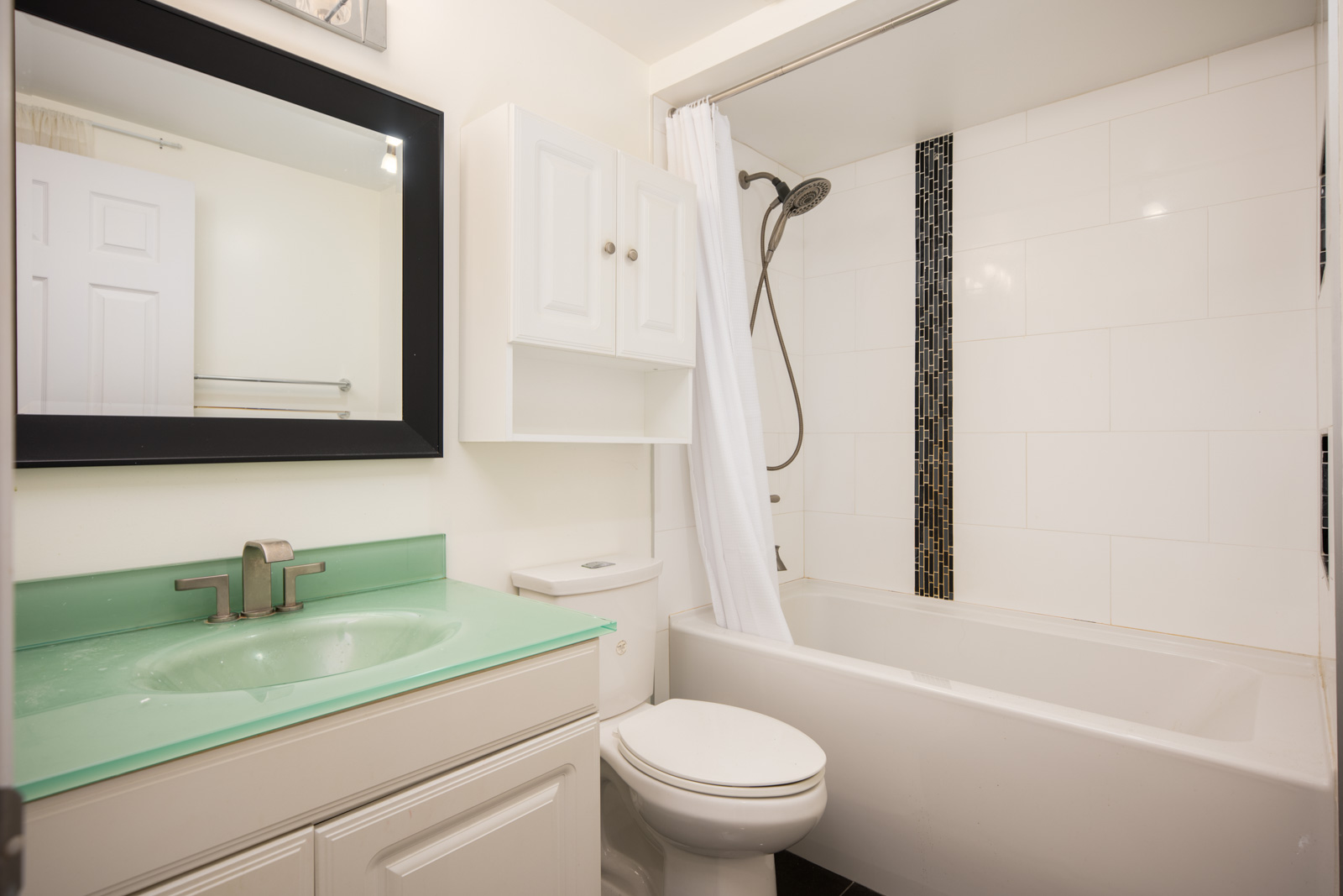 Modern bathroom with a white vanity, green countertop, wall-mounted cabinet, toilet, and bathtub with a shower and white curtain. Vertical tile accent in shower area.
