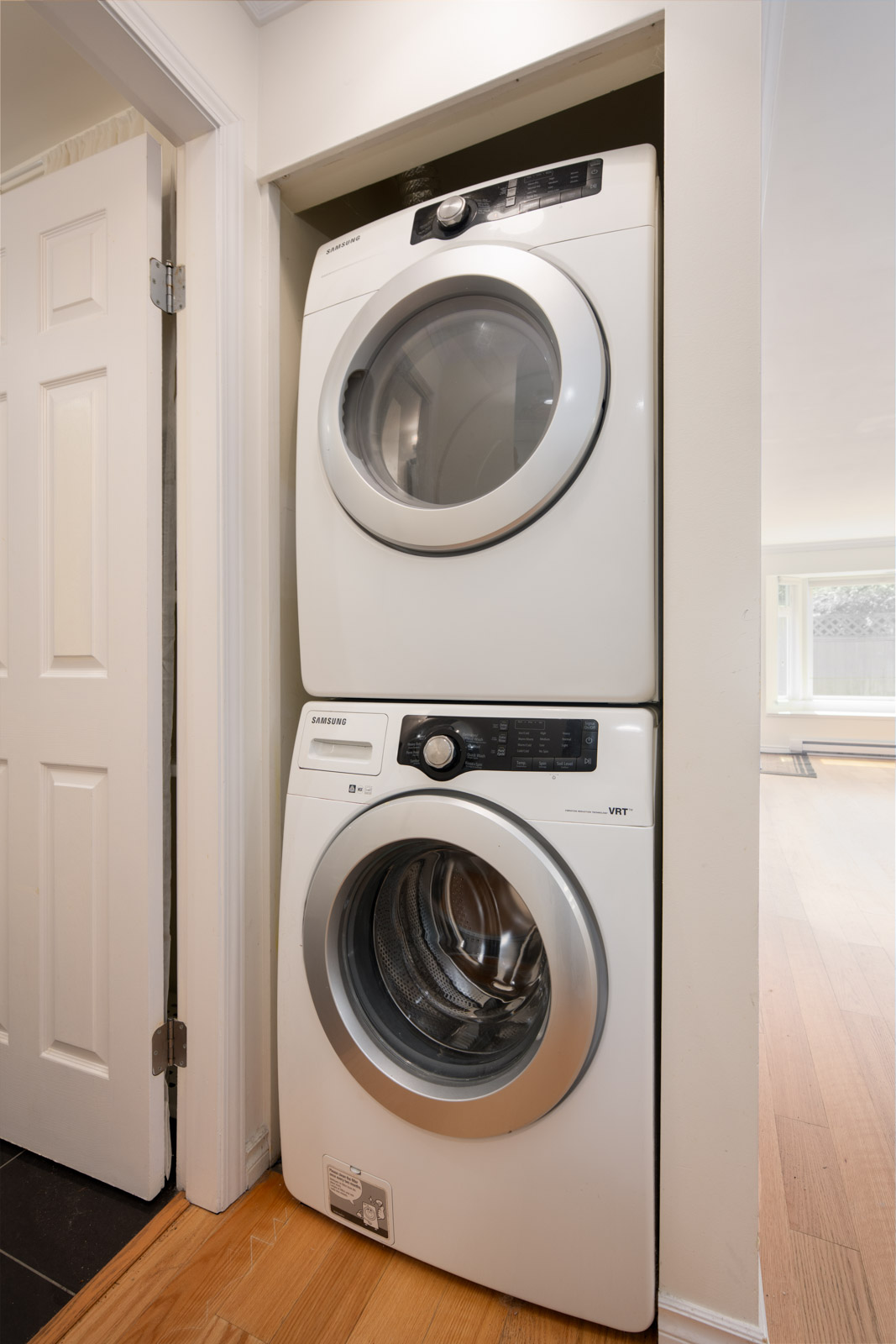 A stacked washer and dryer set installed in a small laundry closet with white walls and a wooden floor.