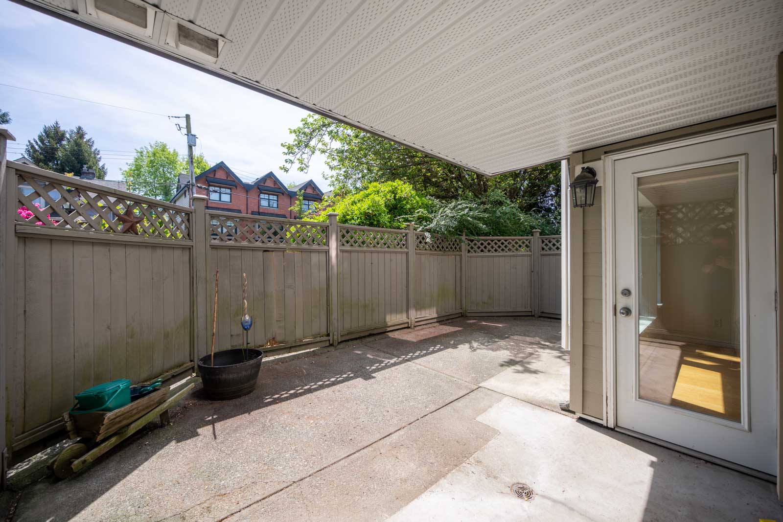 A small, fenced patio area with concrete flooring, a covered section, a glass door leading inside, and some potted plants along the wooden fence.