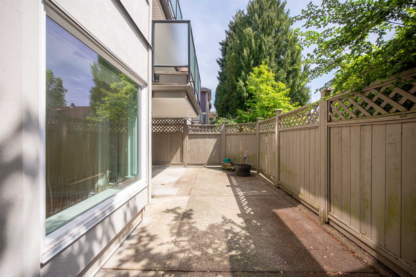 A small, fenced concrete patio beside a modern building, with a few potted plants and trees visible in the background.