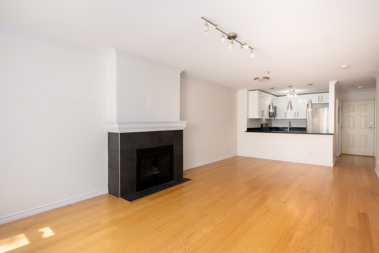 Unfurnished living room with light wood floors, a black tile fireplace, white walls, open kitchen with white cabinets, and stainless steel appliances.