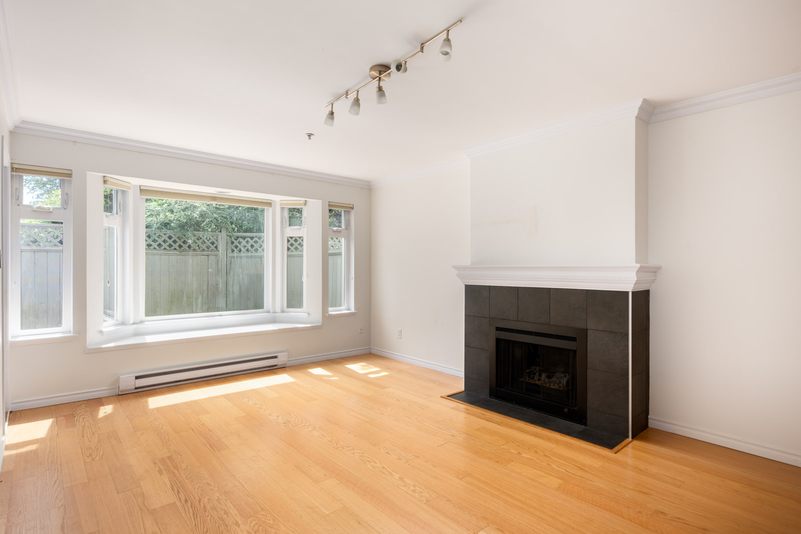 Bright empty living room with light wood floors, a black tile fireplace, large bay window, and white walls.