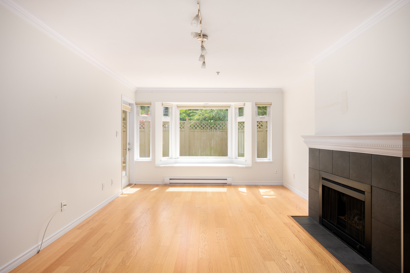 Bright, empty living room with light wood floor, white walls, a black fireplace, and a large bay window overlooking a fenced yard.