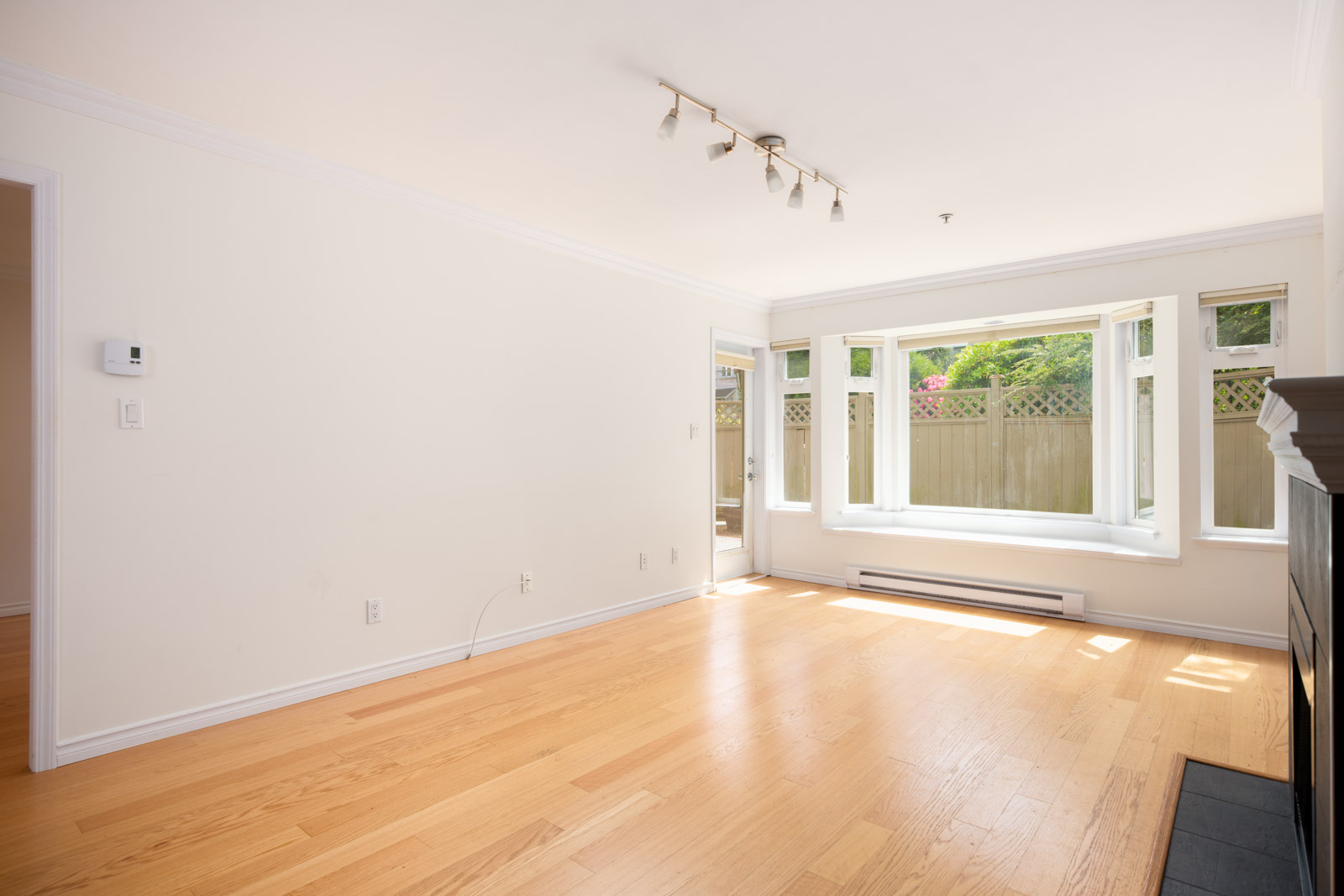 Bright, empty living room with light wood flooring, white walls, a ceiling light fixture, large windows, and a glass door leading outside.