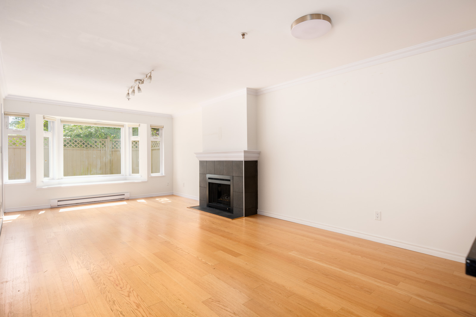 Empty living room with light wood flooring, white walls, large front window, fireplace with black tile surround, and ceiling lights.