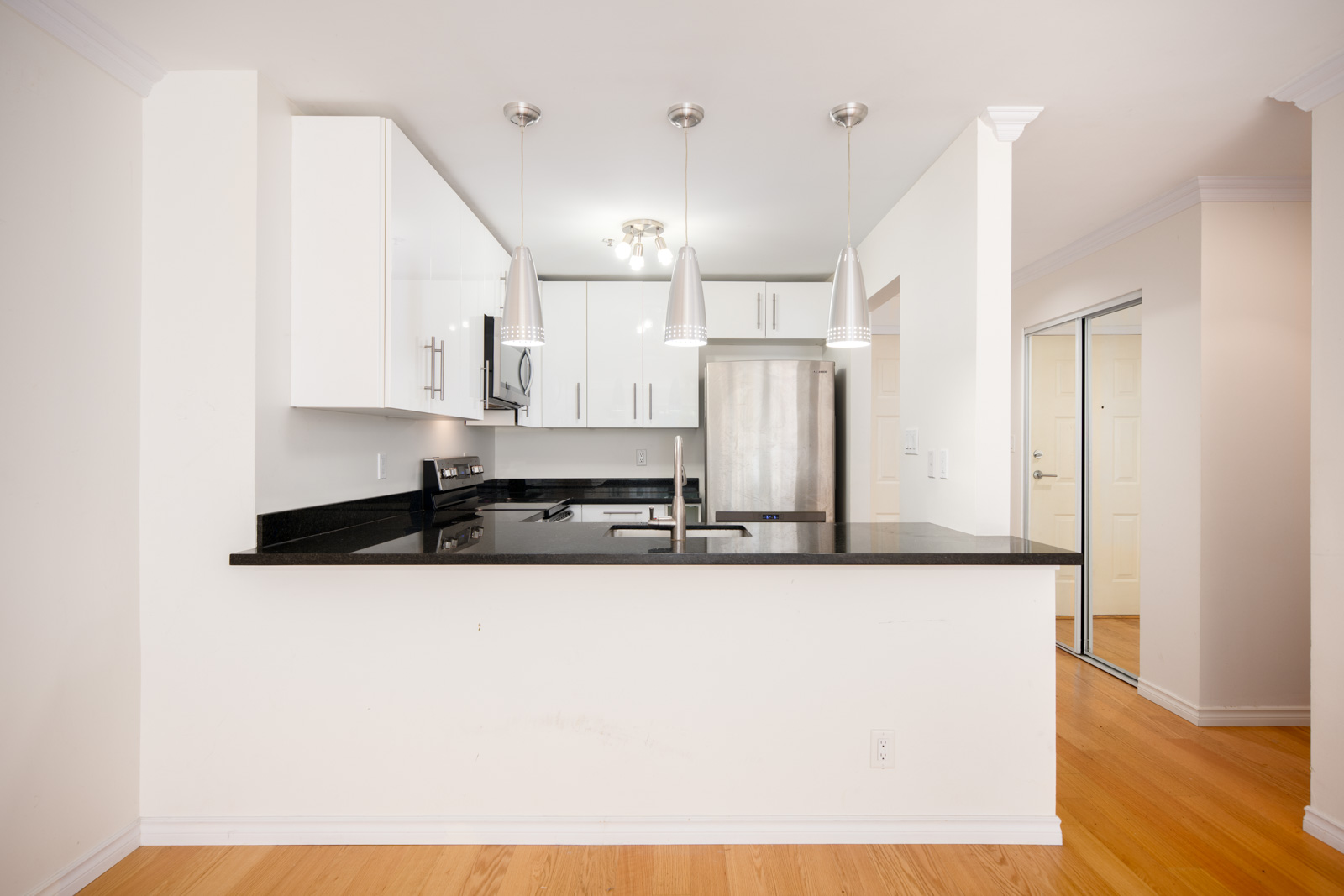 Modern kitchen with white cabinets, stainless steel appliances, three pendant lights, black countertop, and light wood flooring.