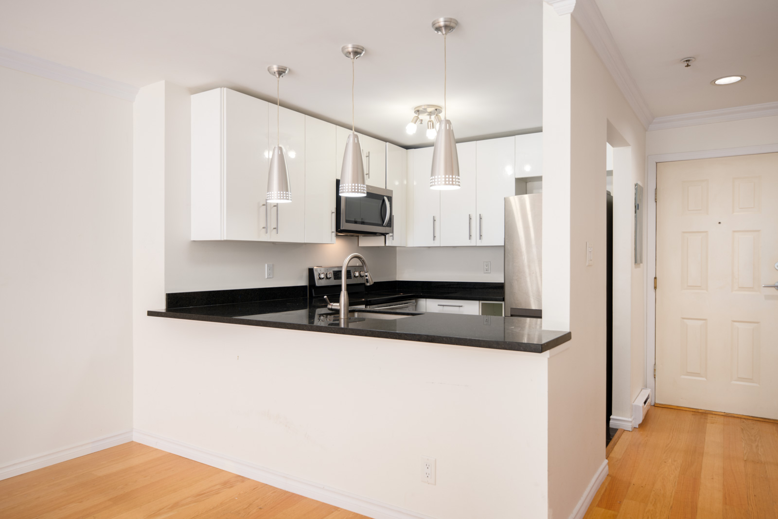 Modern kitchen with white cabinets, black countertops, stainless steel appliances, and three pendant lights above a peninsula counter; light wood flooring and neutral walls.