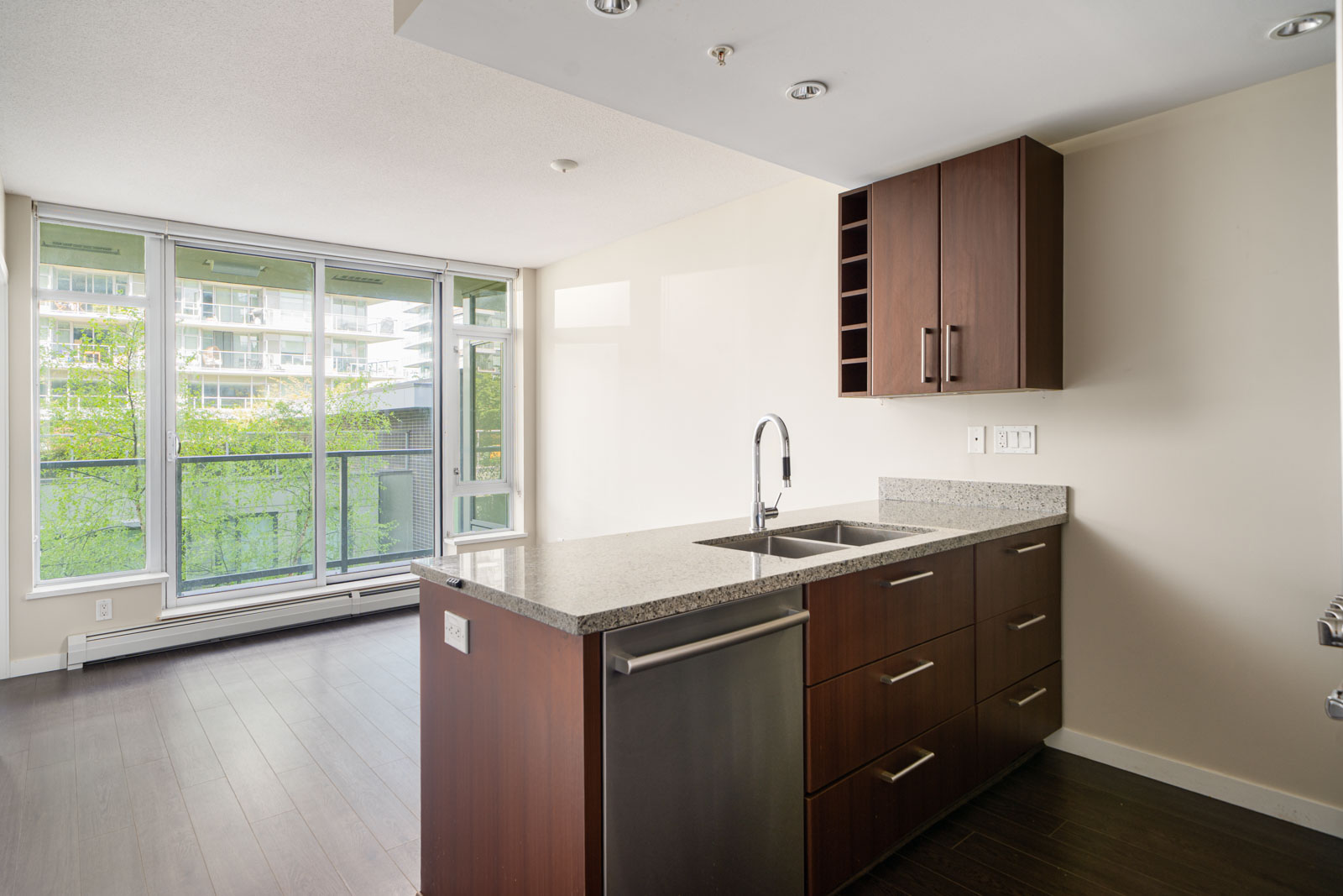 Modern apartment kitchen with a center island, built-in sink, dark wood cabinets, and large windows overlooking a balcony and green trees.