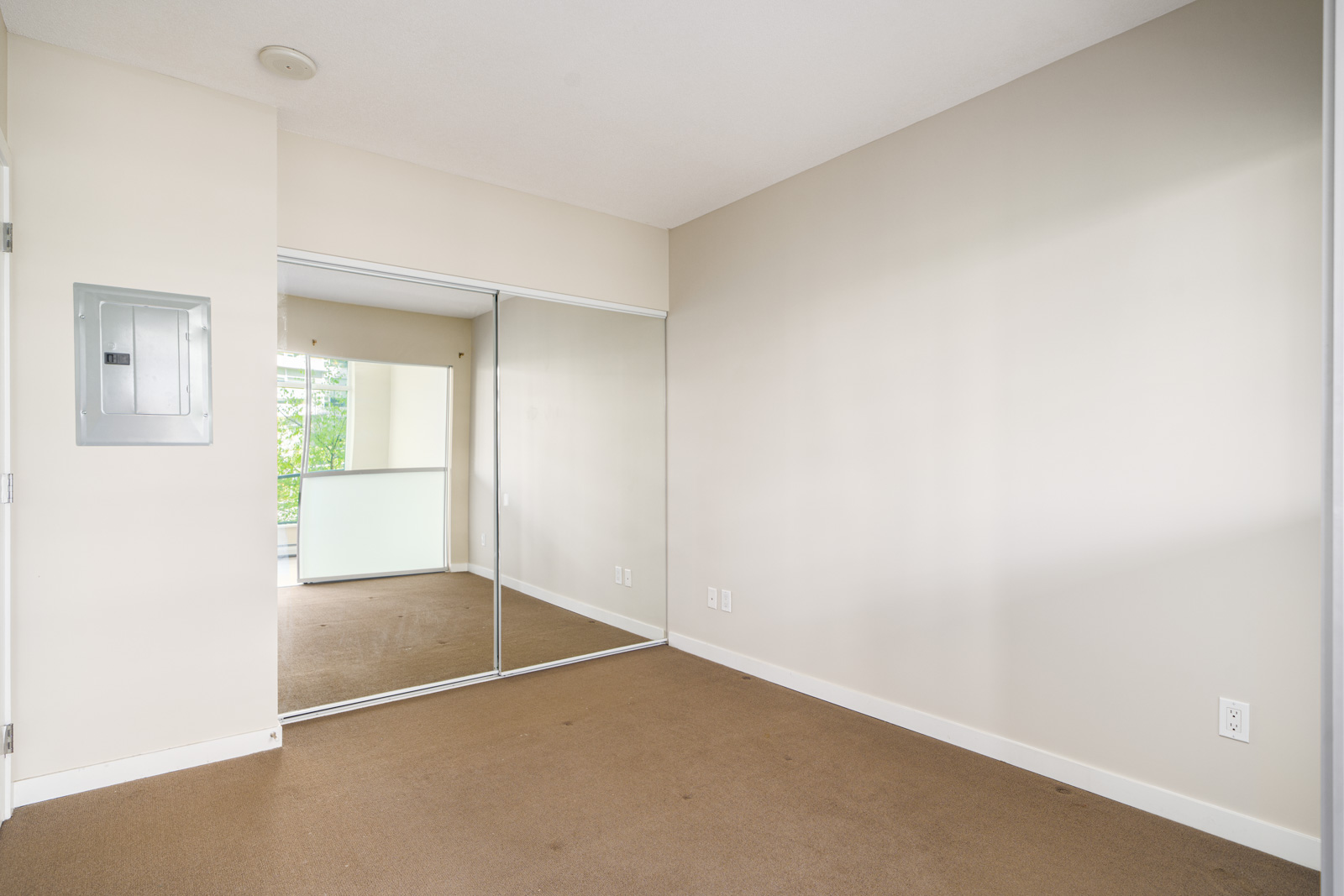 Empty room with beige carpet, off-white walls, a mirrored sliding closet door, and an electrical panel on the left wall. Natural light coming from a window reflected in the mirror.