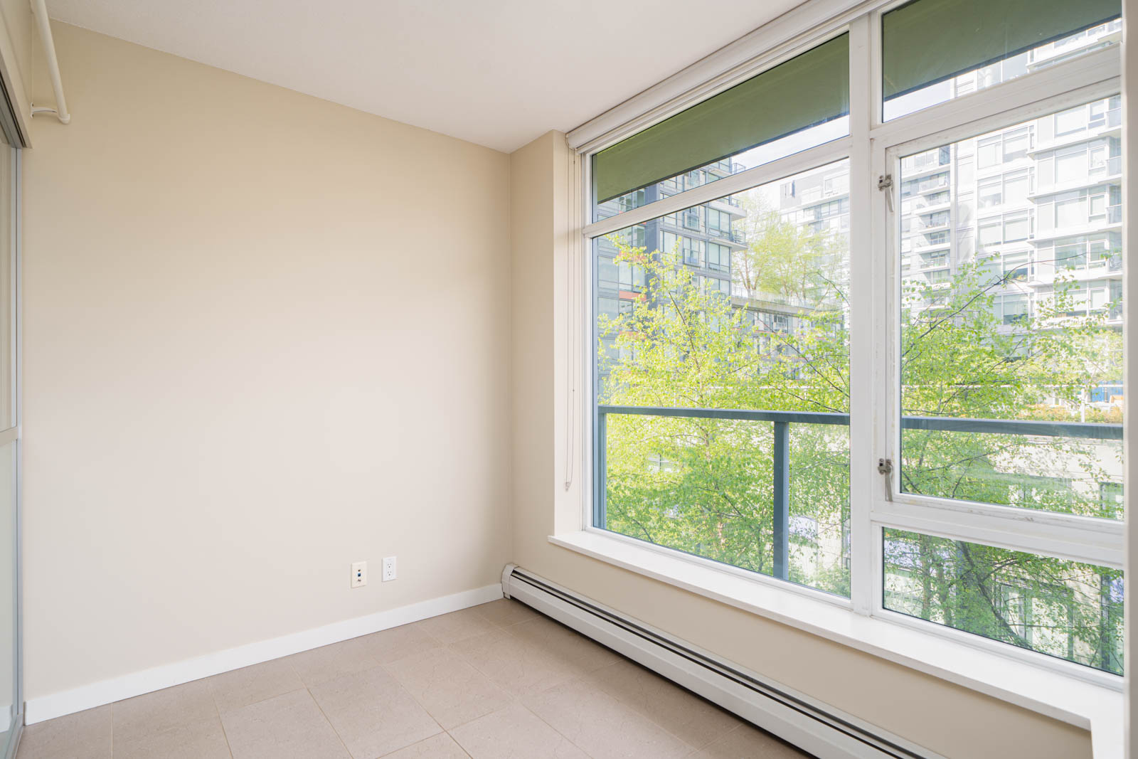 Empty room with beige walls, large windows overlooking trees and nearby buildings, tiled floor, and baseboard heater.