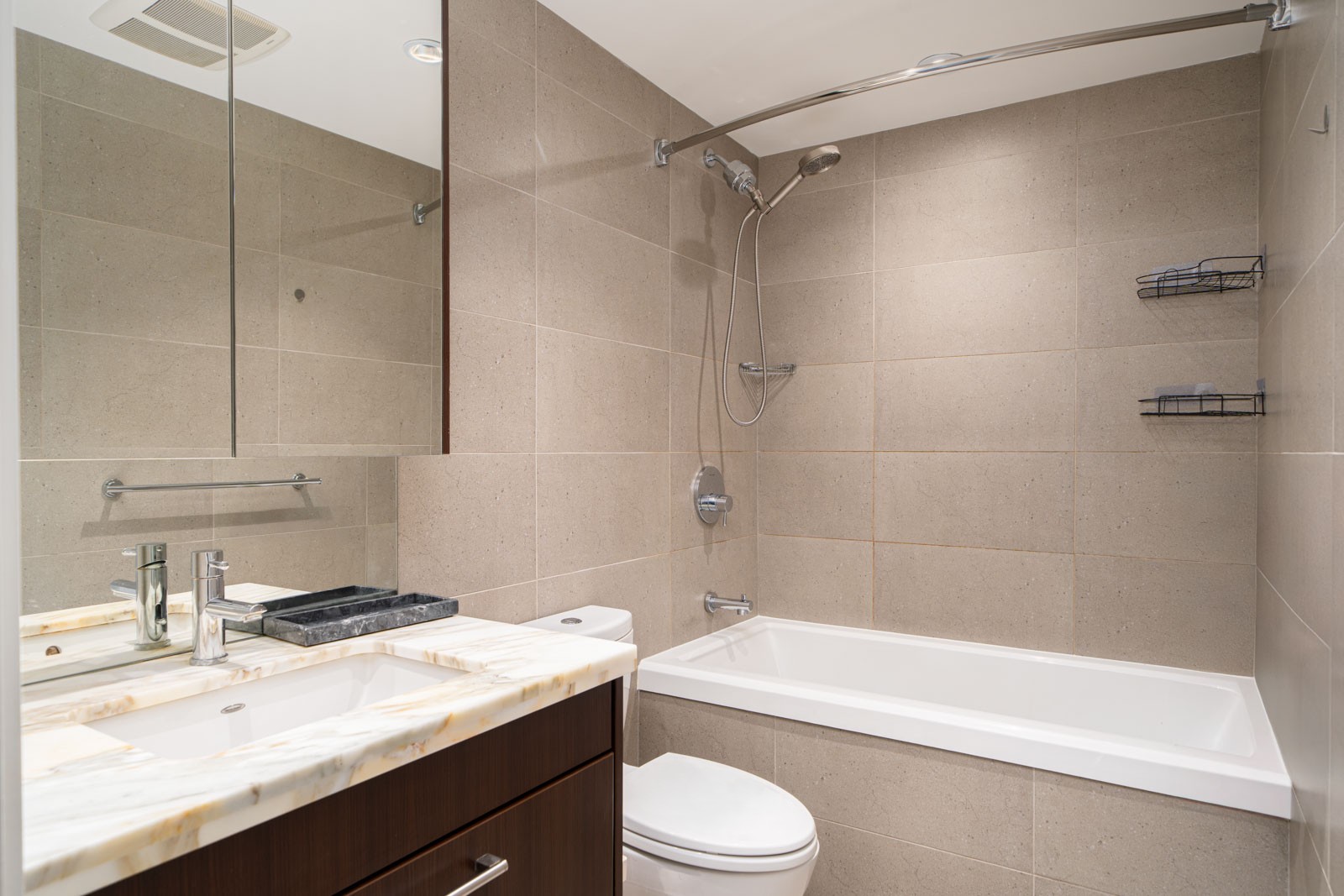 Modern bathroom with a marble countertop, under-mount sink, wall-mounted mirror, toilet, and a bathtub with a showerhead and wire racks on tiled walls.