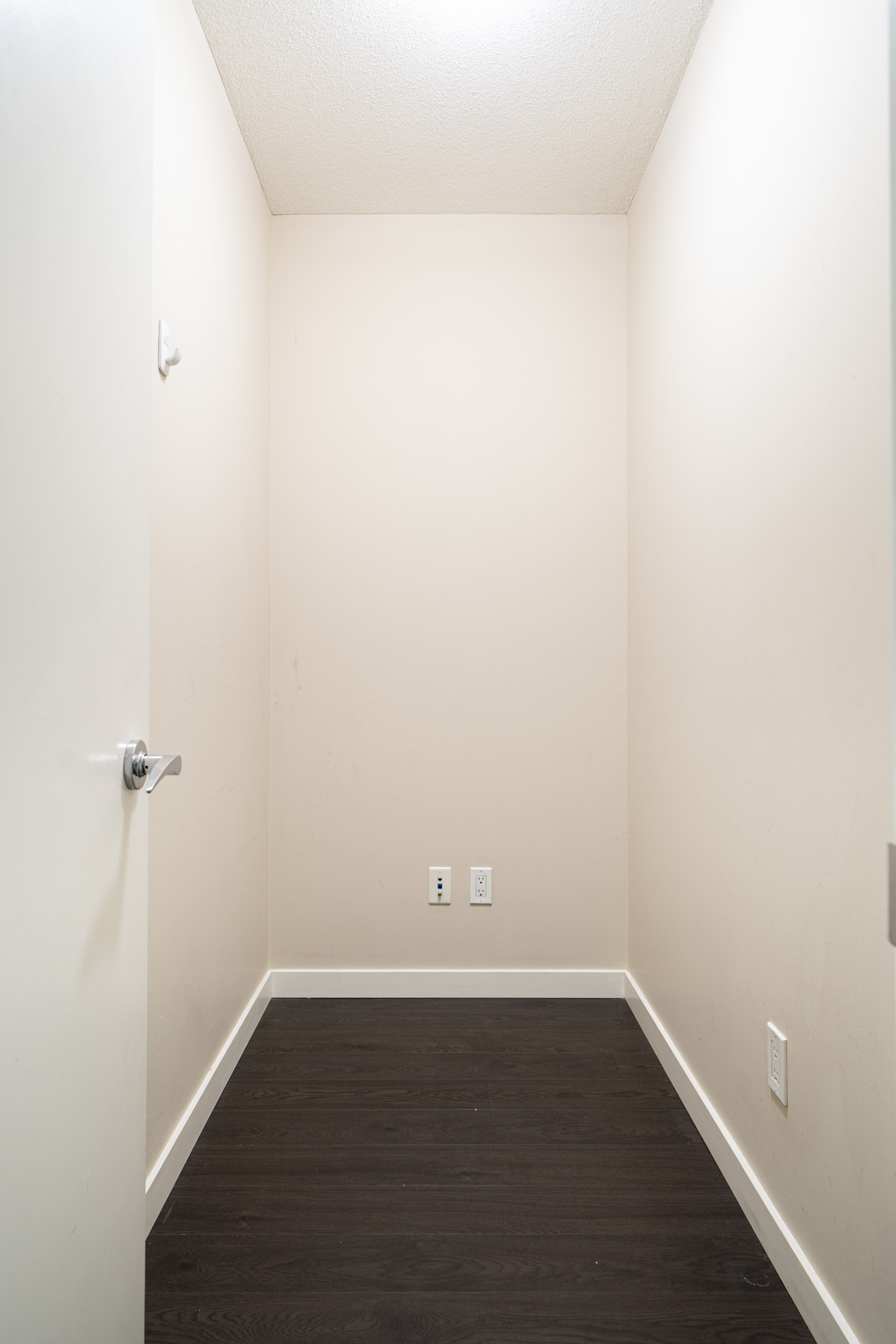 An empty, small rectangular room with cream-colored walls, dark wood flooring, a partially open door, and electrical outlets on one wall.