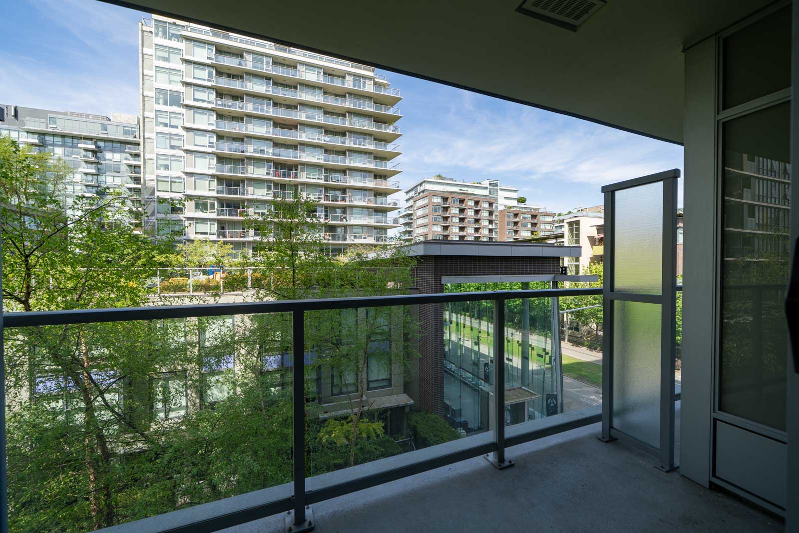 View from a modern balcony with glass railing overlooking trees and several mid-rise apartment buildings on a sunny day.