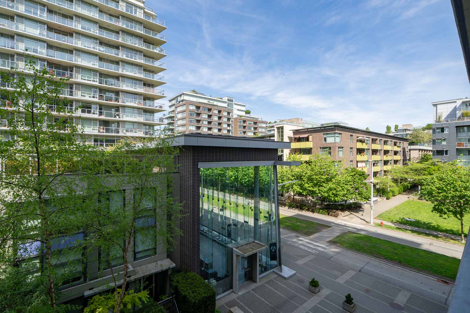 View of modern apartment buildings and a glass entrance structure overlooking a landscaped courtyard and tree-lined walkway on a sunny day.