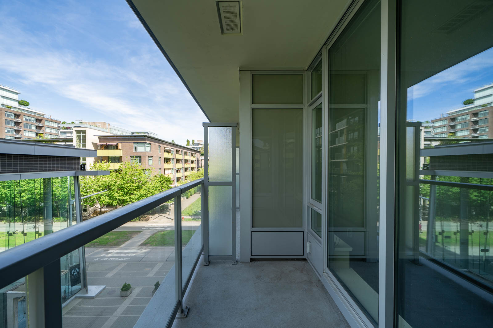 A modern apartment balcony with glass railings and large windows, overlooking a courtyard with green trees and nearby residential buildings under a blue sky.