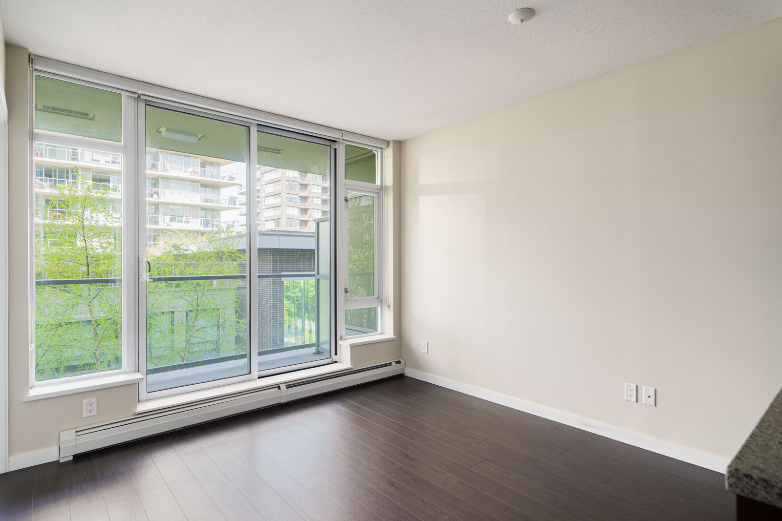 Empty room with dark wood flooring, large floor-to-ceiling windows, sliding glass door to a balcony, and light-colored walls, overlooking trees and nearby buildings.