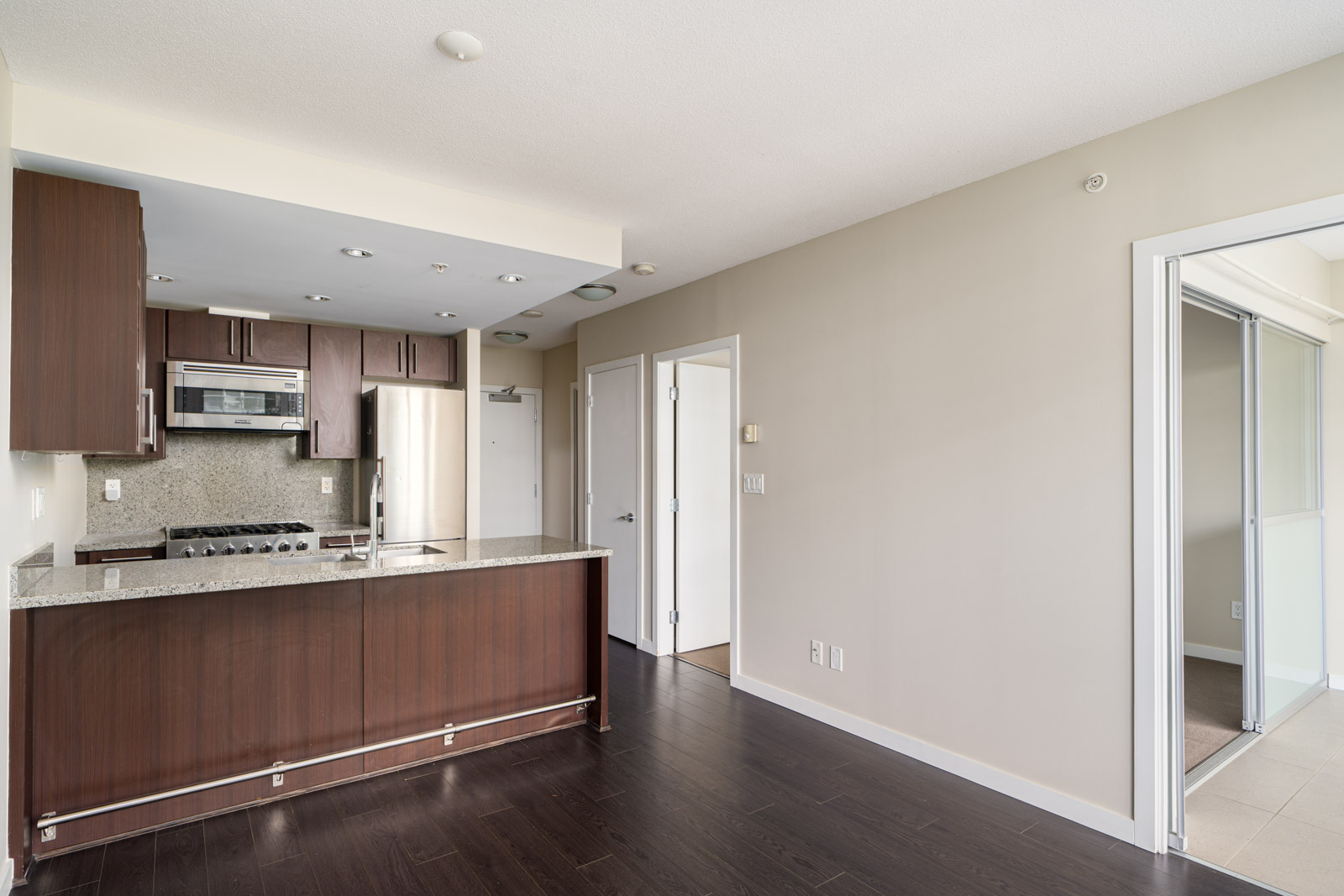 Modern kitchen with dark wood cabinets, stainless steel appliances, and granite countertops adjacent to an empty room with sliding glass doors.