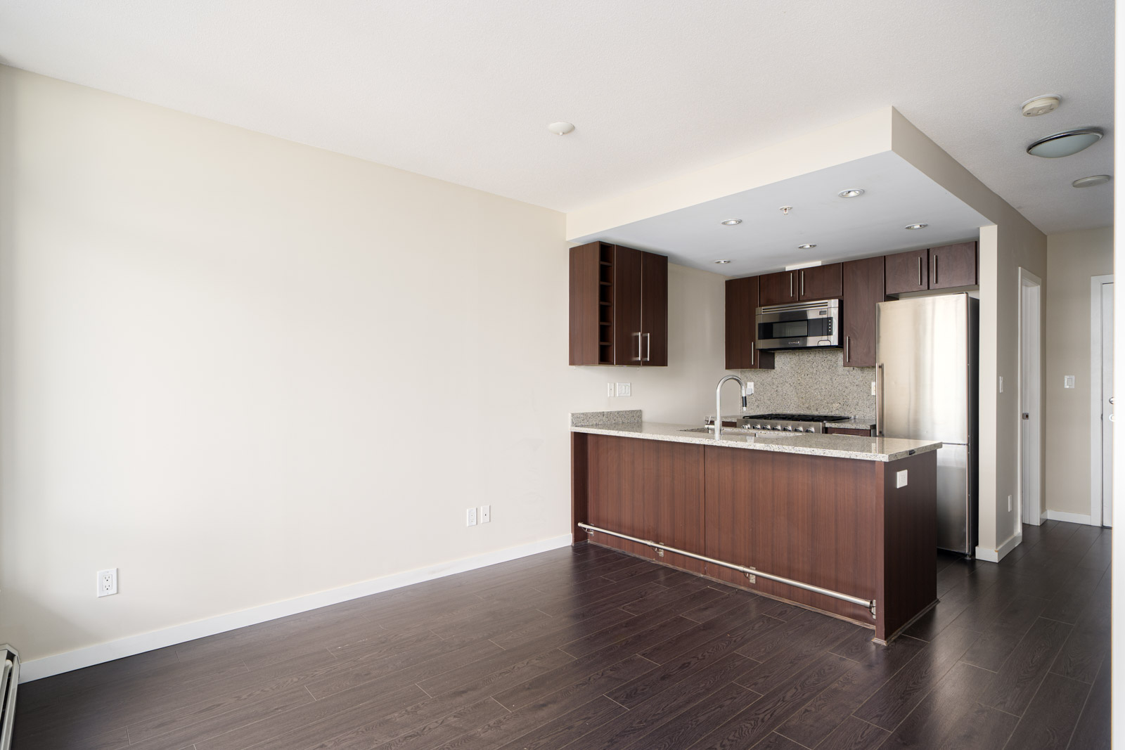 Modern kitchen with dark wood cabinets, granite countertops, stainless steel appliances, and open layout connected to a bright living area with light-colored walls and dark hardwood floors.