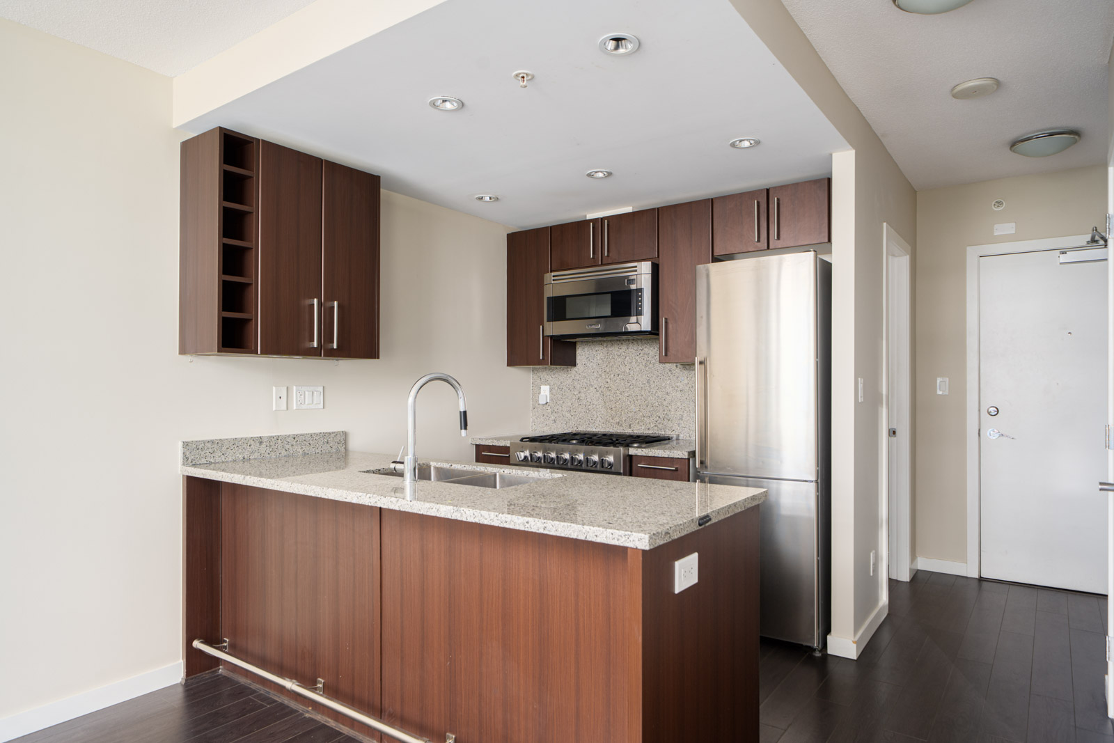 Modern kitchen with dark wood cabinets, stainless steel appliances, granite countertops, and an island with a sink. Walls are neutral-colored and lighting is recessed.