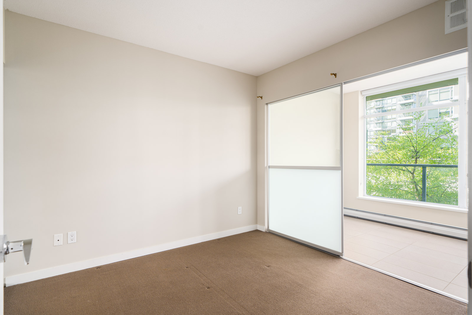Empty room with beige walls and brown carpet, adjoining a bright balcony area through a sliding glass door; large window with view of green trees and buildings outside.